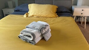 Un lit bien fait, avec une couverture jaune, une serviette pliée aux motifs gris et blancs, et un oreiller bleu.