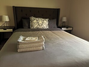 Een net opgemaakt bed met een opgevouwen handdoek en een decoratief kussen, afgezet door twee nachtkastjes met lampen.