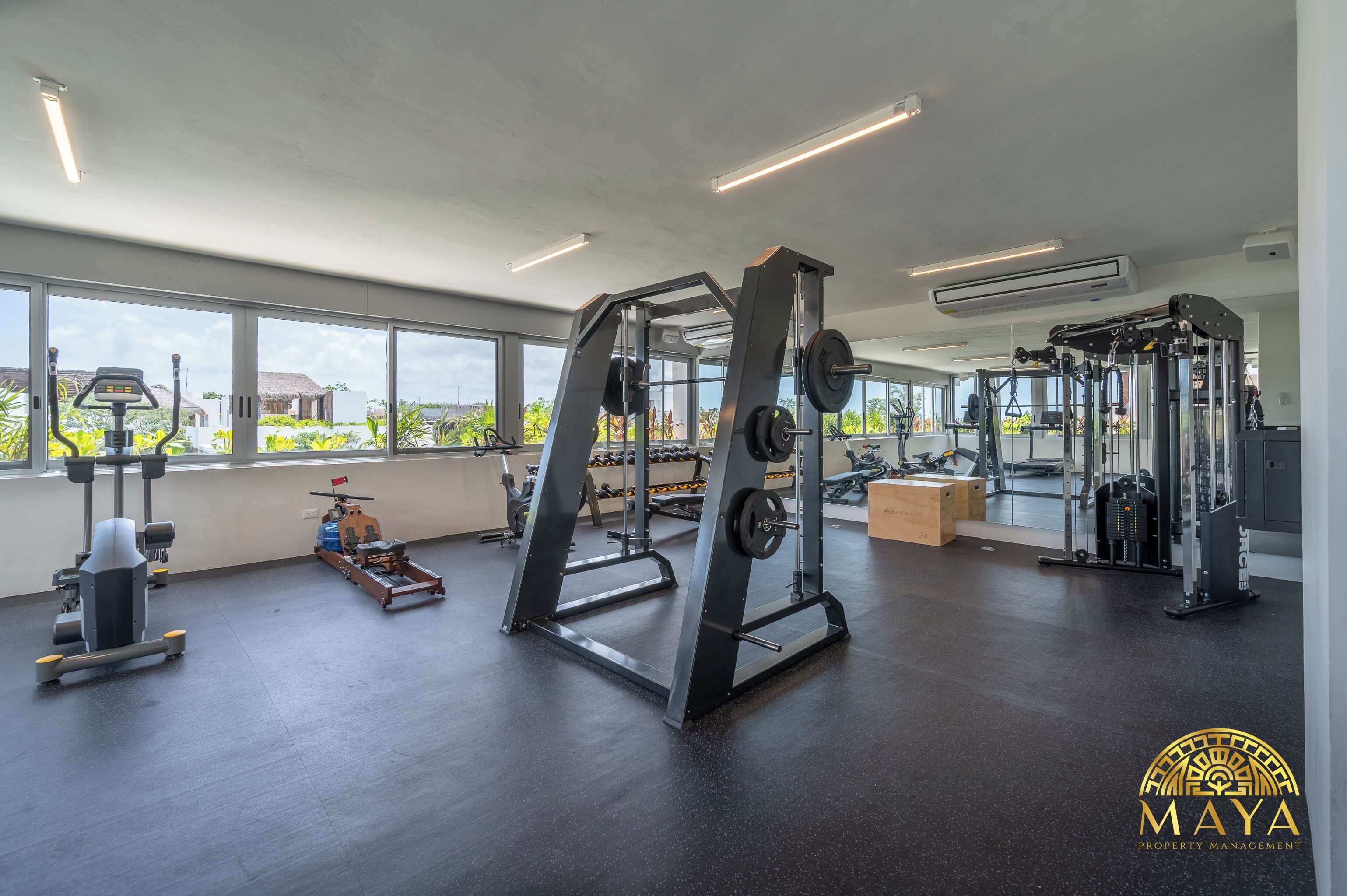 Een sportschool met diverse trainingsapparaten, grote ramen en uitzicht op groen.