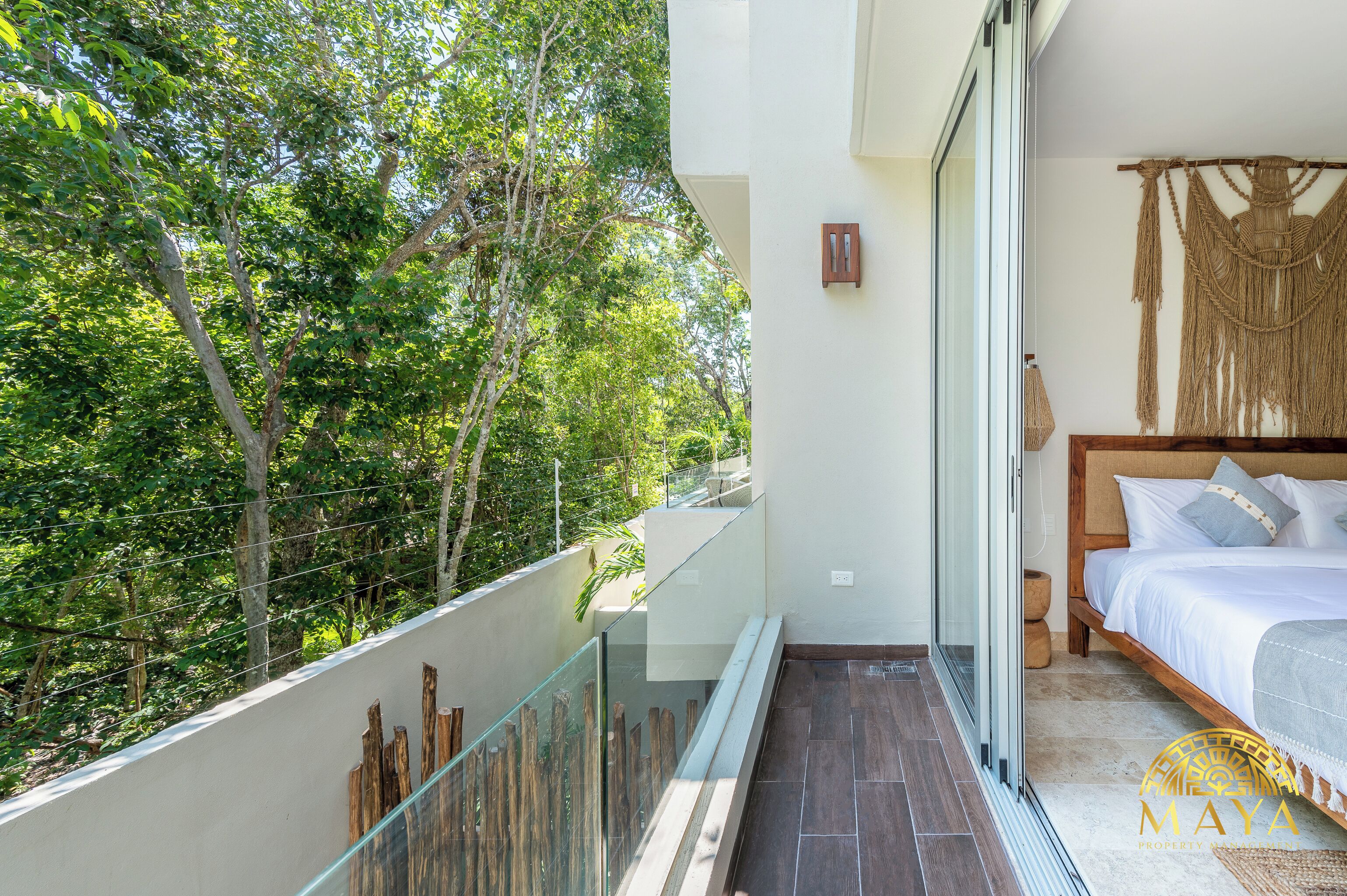 Un balcon doté d’une balustrade en verre donnant sur un espace verdoyant luxuriant, un lit avec une tête de lit en bois et une décoration murale en macramé.