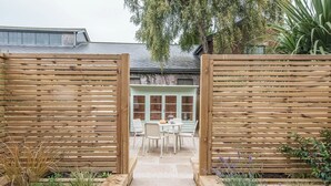 Une porte en bois mène à un espace patio aménagé avec une table et des chaises, le tout entouré de parterres surélevés où poussent des plantes.