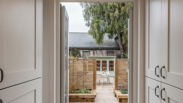 On aperçoit, à travers une porte ouverte, une cour intérieure avec une table et des chaises, une clôture en bois et, au fond, un bâtiment dont le toit est en pente.