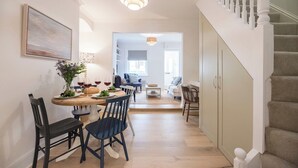 Un coin repas avec une table ronde, des chaises et un vase de fleurs. Un escalier se trouve sur la droite, et on aperçoit une autre pièce avec un canapé et une fenêtre.