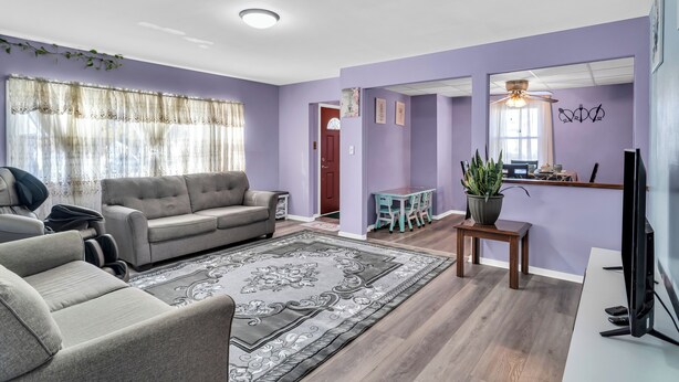 A living room with a grey sofa, a coffee table, and a TV. There is a dining area with a table and chairs, and a kitchen area with a counter and a plant.