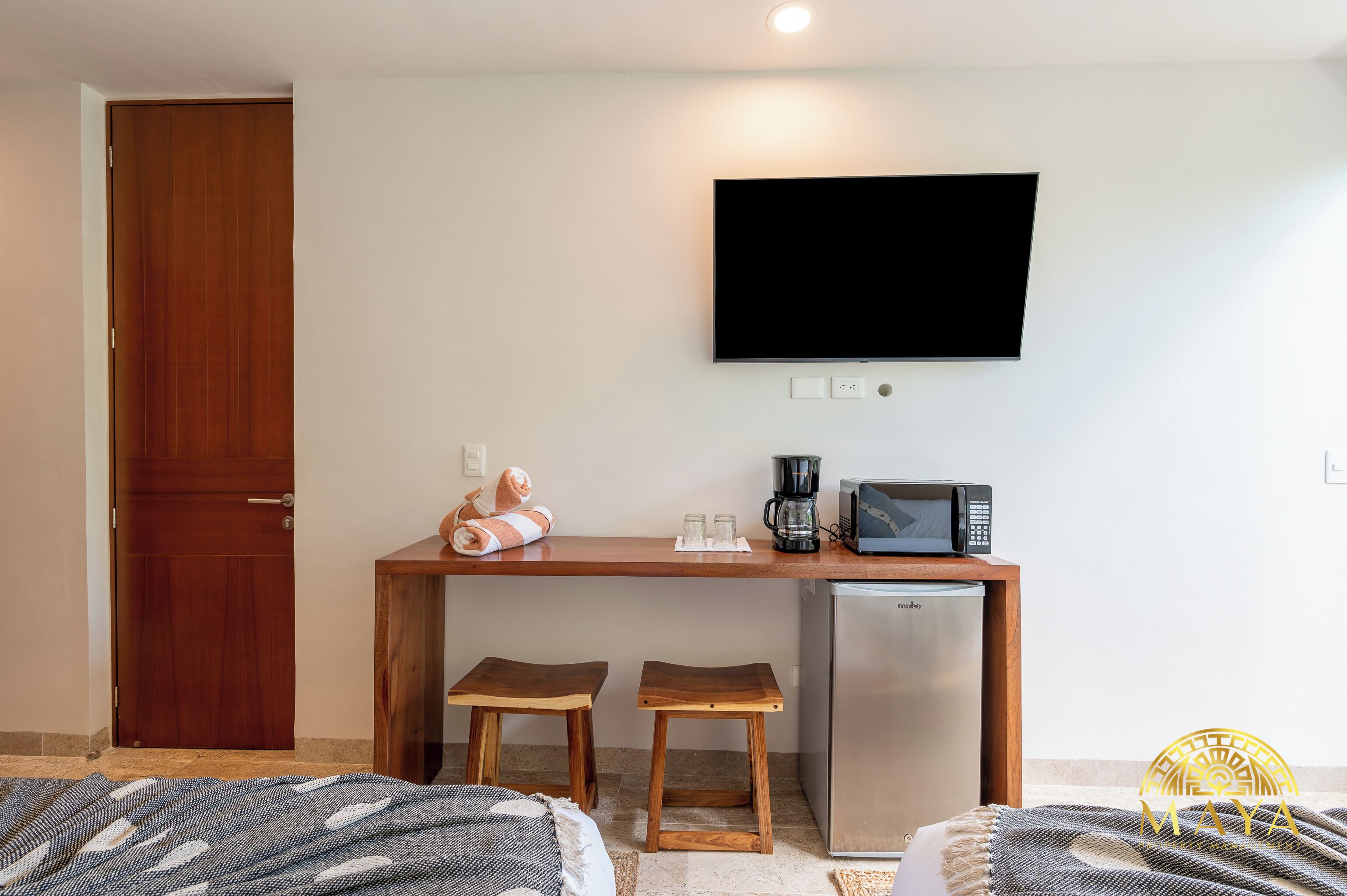 A compact room with a wooden desk, two stools, a flat-screen TV, a coffee maker, and a mini-fridge.
