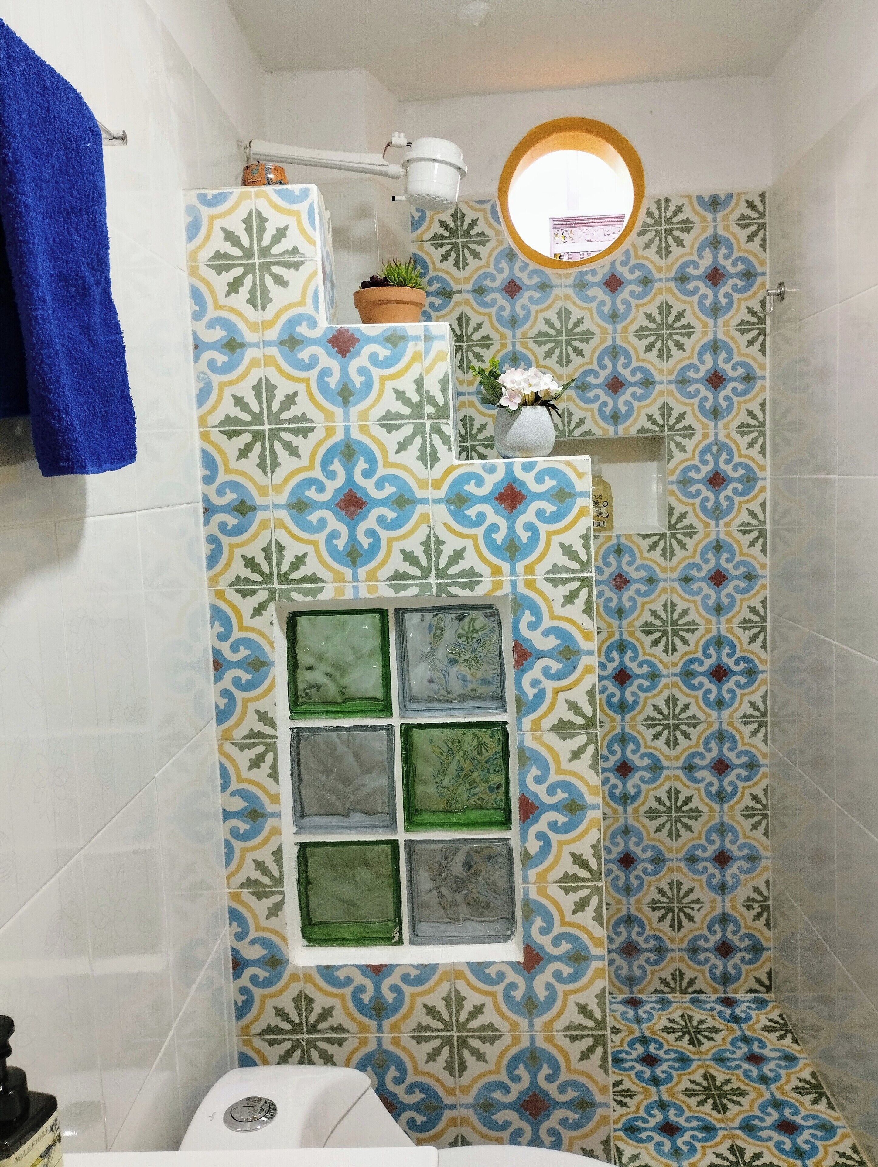 Casa de banho com azulejos estampados, toalha azul, pequena janela e planta em vaso.