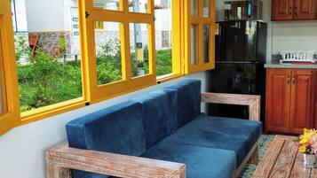 A room with a blue sofa, wooden-framed chairs, a kitchen with wooden cabinets, and a large window with yellow frames.