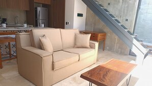 A modern living room with a beige sofa, a wooden coffee table, and a kitchen area with wooden cabinets and a refrigerator. - New, Modern Loft in Main Avenue Tulum (Tulum)