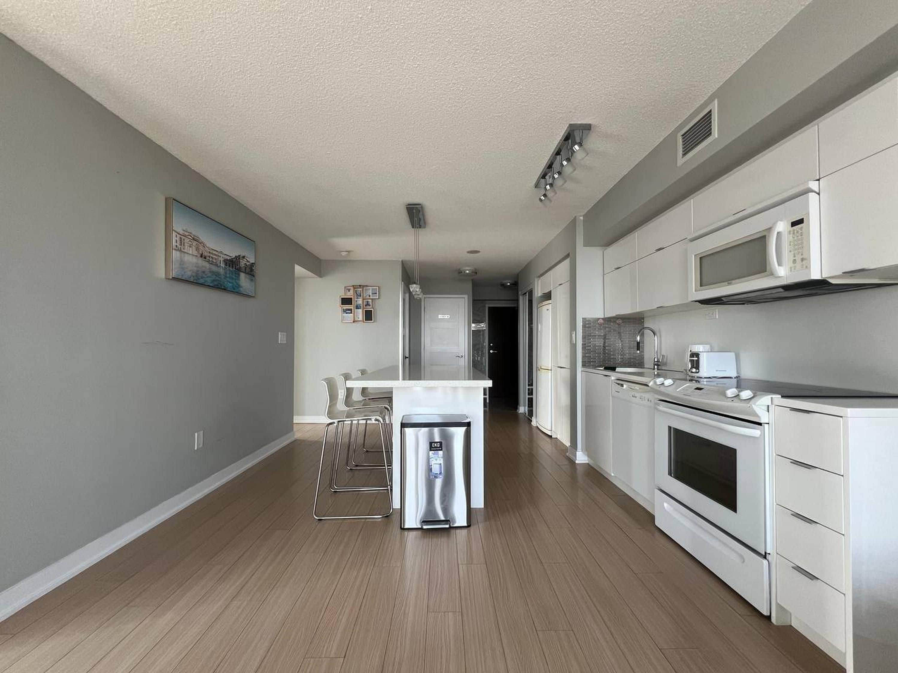A modern kitchen with stainless steel appliances, a dining area with bar stools, and a hallway leading to a door.