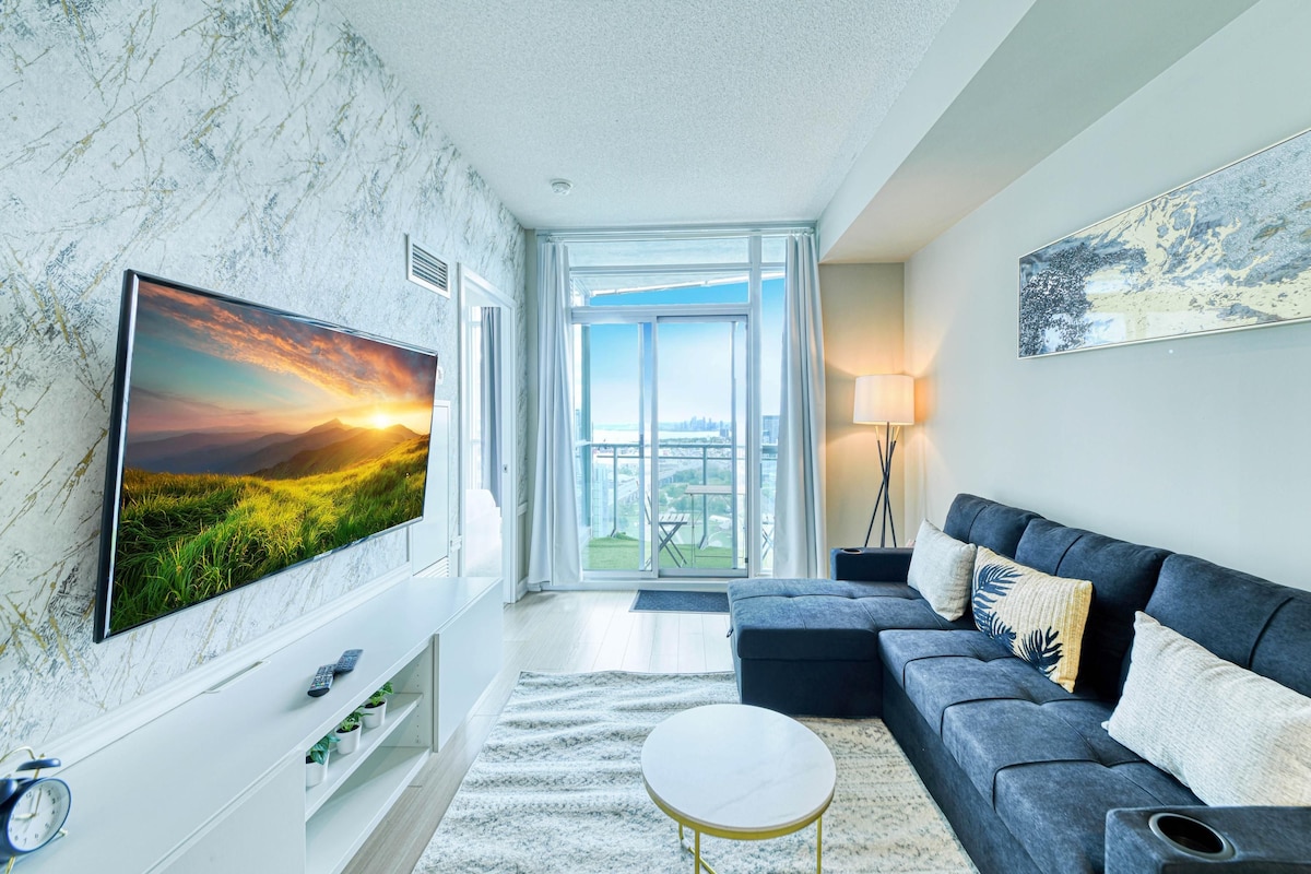 A modern living room with a large flat-screen TV, a dark blue sofa, a round coffee table, and a view of a cityscape through large windows.