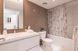 Un baño moderno con un lavamanos blanco, un inodoro y una bañera. Hay una cortina de ducha con un motivo de botellas y una planta en maceta sobre la bañera.