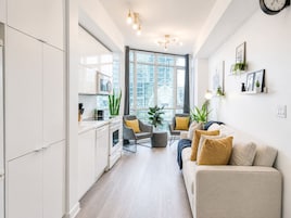 Cocina moderna con armarios blancos, microondas y estufa. Hay un área de comedor con sofá gris y cojines amarillos. La habitación tiene ventanas amplias y vistas a edificios.