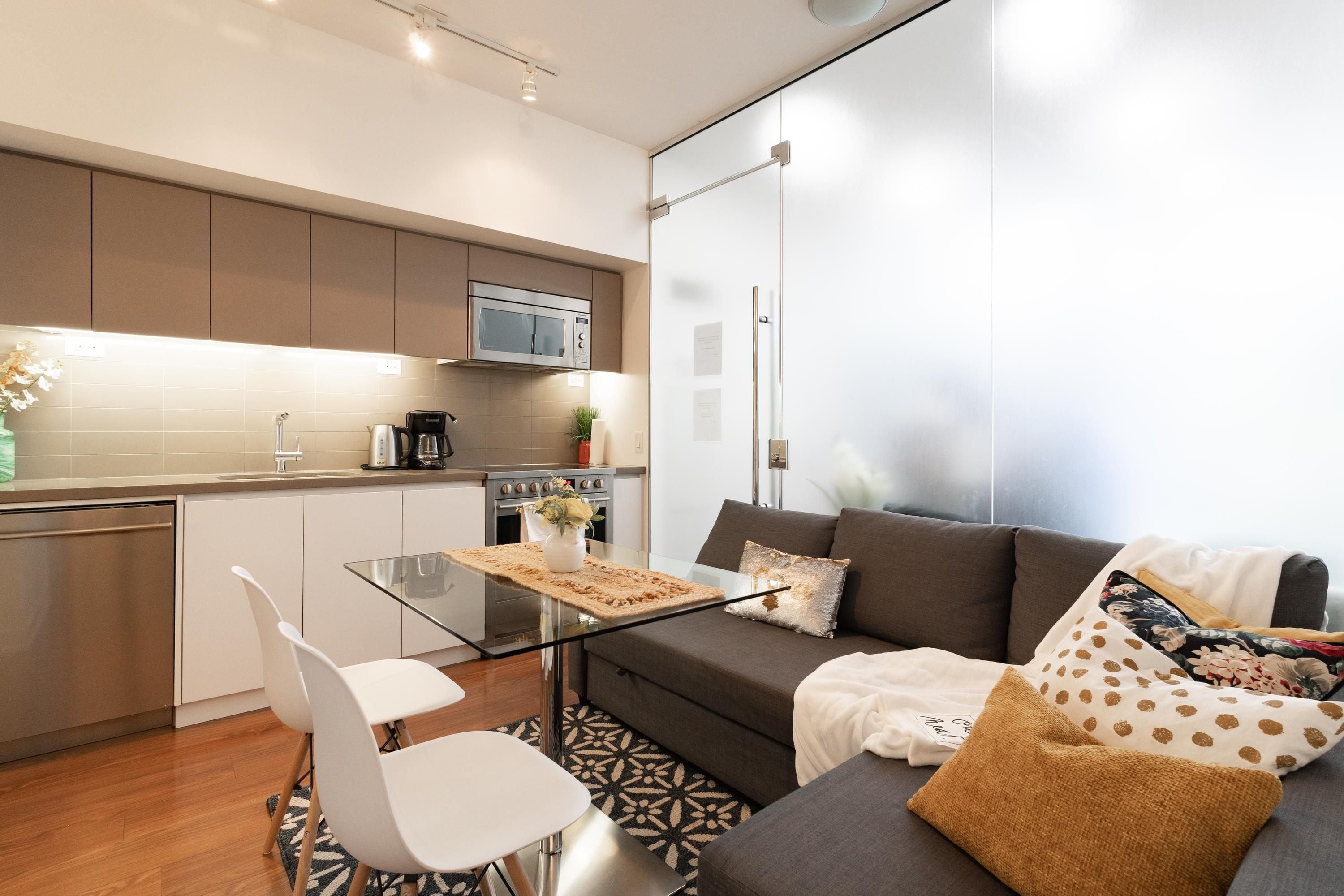 Cocina moderna con microondas, estufa y cafetera. Hay una mesa de vidrio con un mantel individual y una silla blanca. En la sala de estar, un sofá gris con cojines estampados.