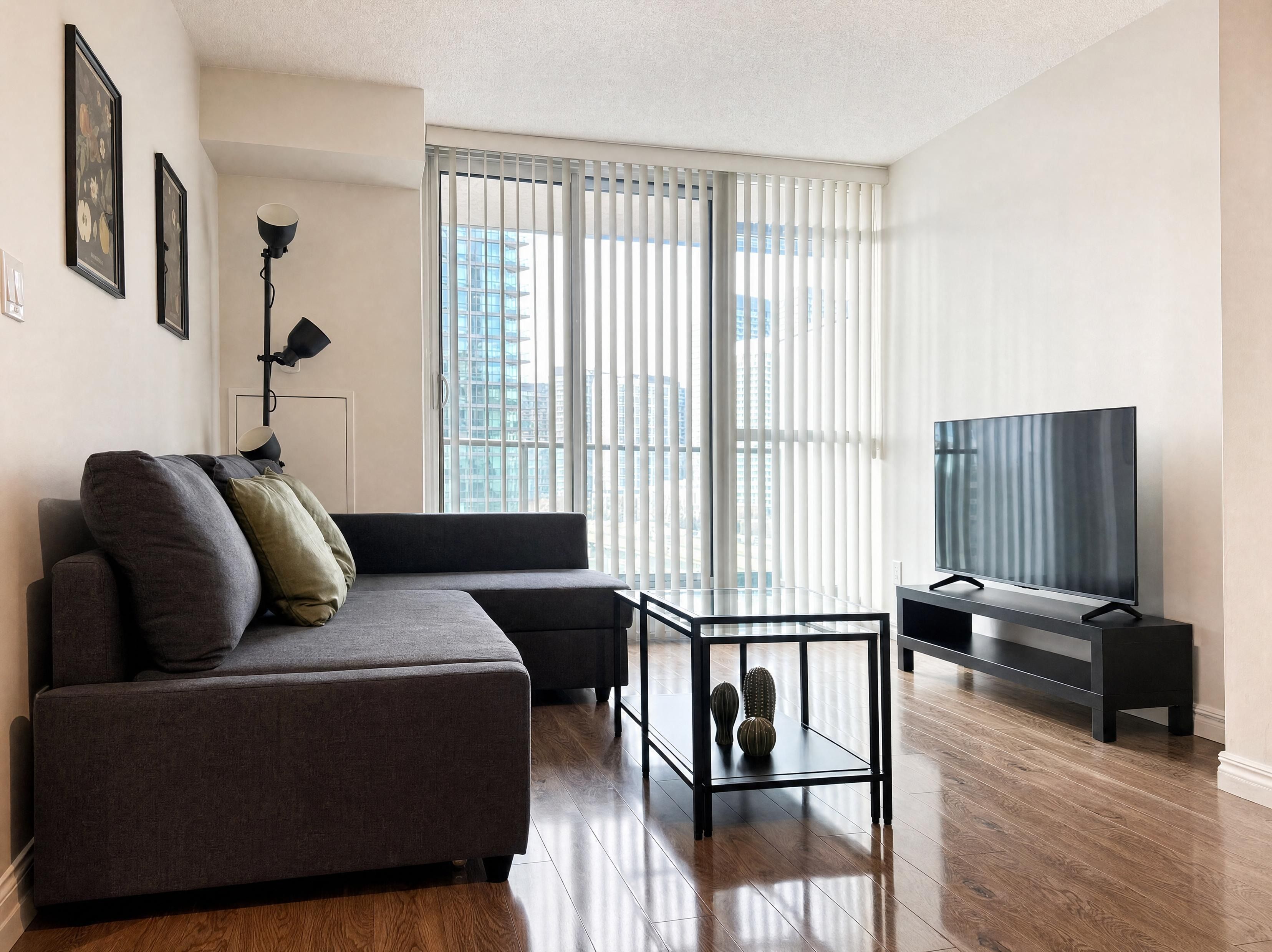 A modern living room with a sofa, a glass coffee table, a flat-screen TV, and a large window with blinds.