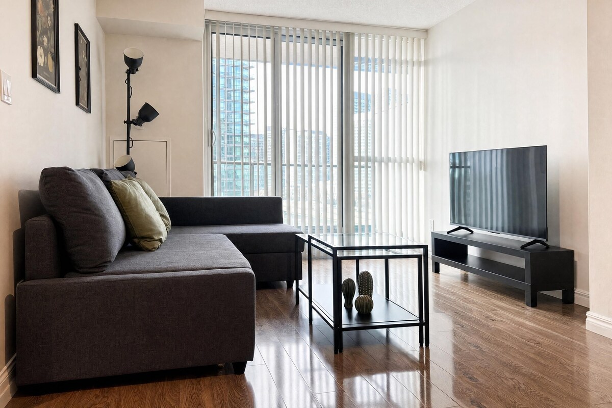 A modern living room with a sofa, a glass coffee table, a flat-screen TV, and a large window with blinds.