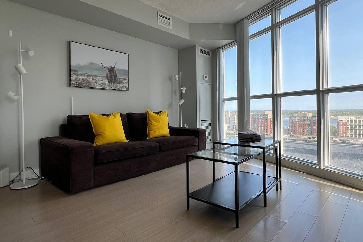 A modern living room with a dark brown sofa, two yellow cushions, a glass coffee table, and a large window offering a city view.