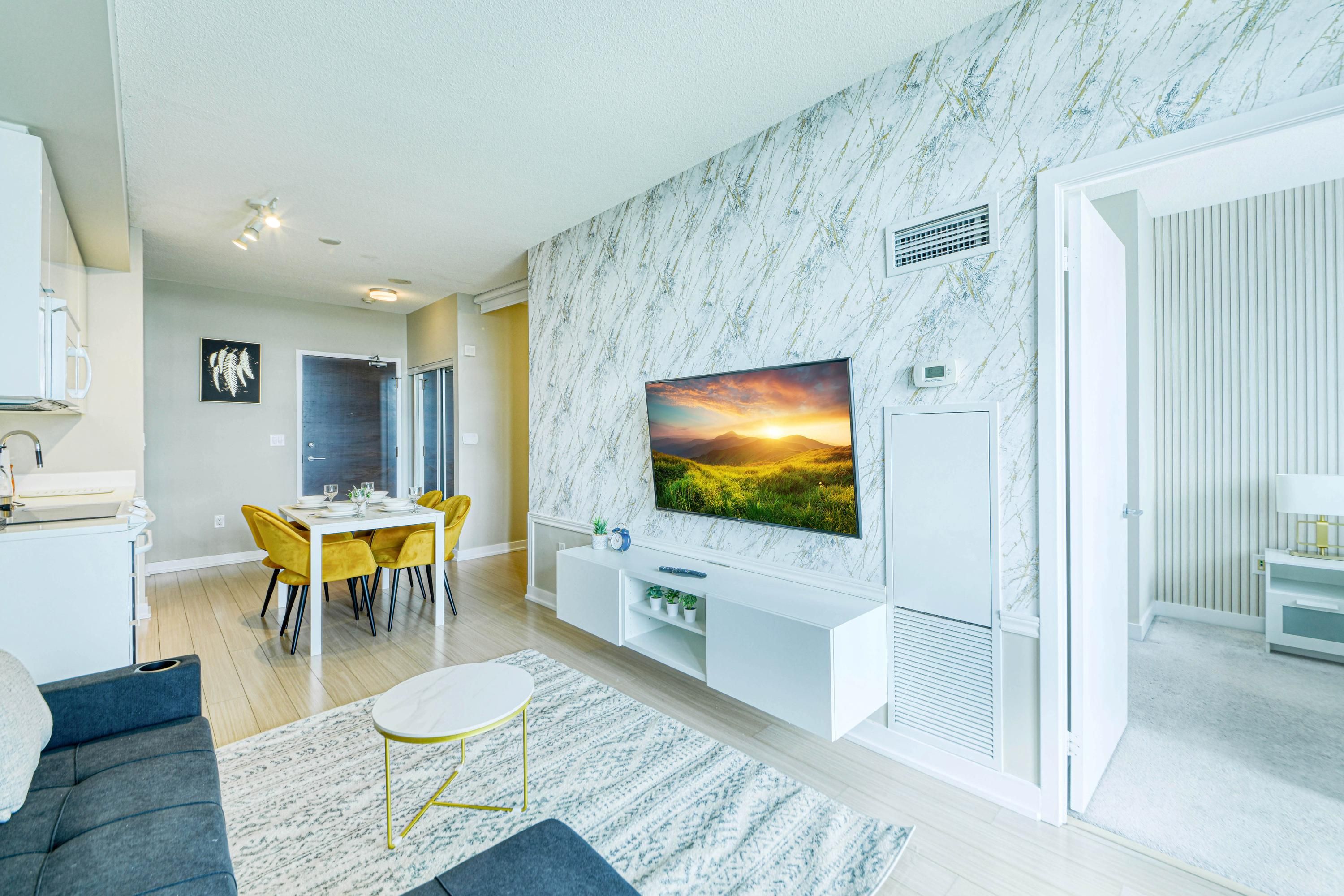 A modern living room with a large TV, a dining area with yellow chairs, and a kitchen area with white cabinets.