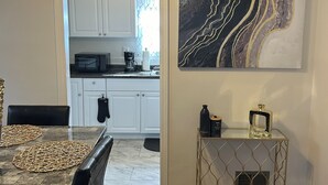 Uma área de refeições com mesa, cadeiras e ventilador de teto. Ao fundo, cozinha com armários brancos e micro-ondas. Um console de metal decorativo está encostado na parede, com uma obra de arte semelhante a mármore sobre ele.