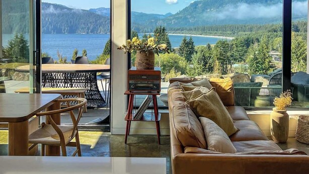 Un salon moderne doté de grandes fenêtres, d’un plafond en bois et offrant une vue sur les montagnes et un lac.