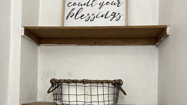 A corner with a wooden shelf and a wire basket. Above the shelf, there is a framed sign with the text "count your blessings".