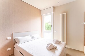 Une chambre d’hôtel moderne avec un grand lit, une fenêtre avec des stores, un radiateur et une vue sur la verdure environnante.