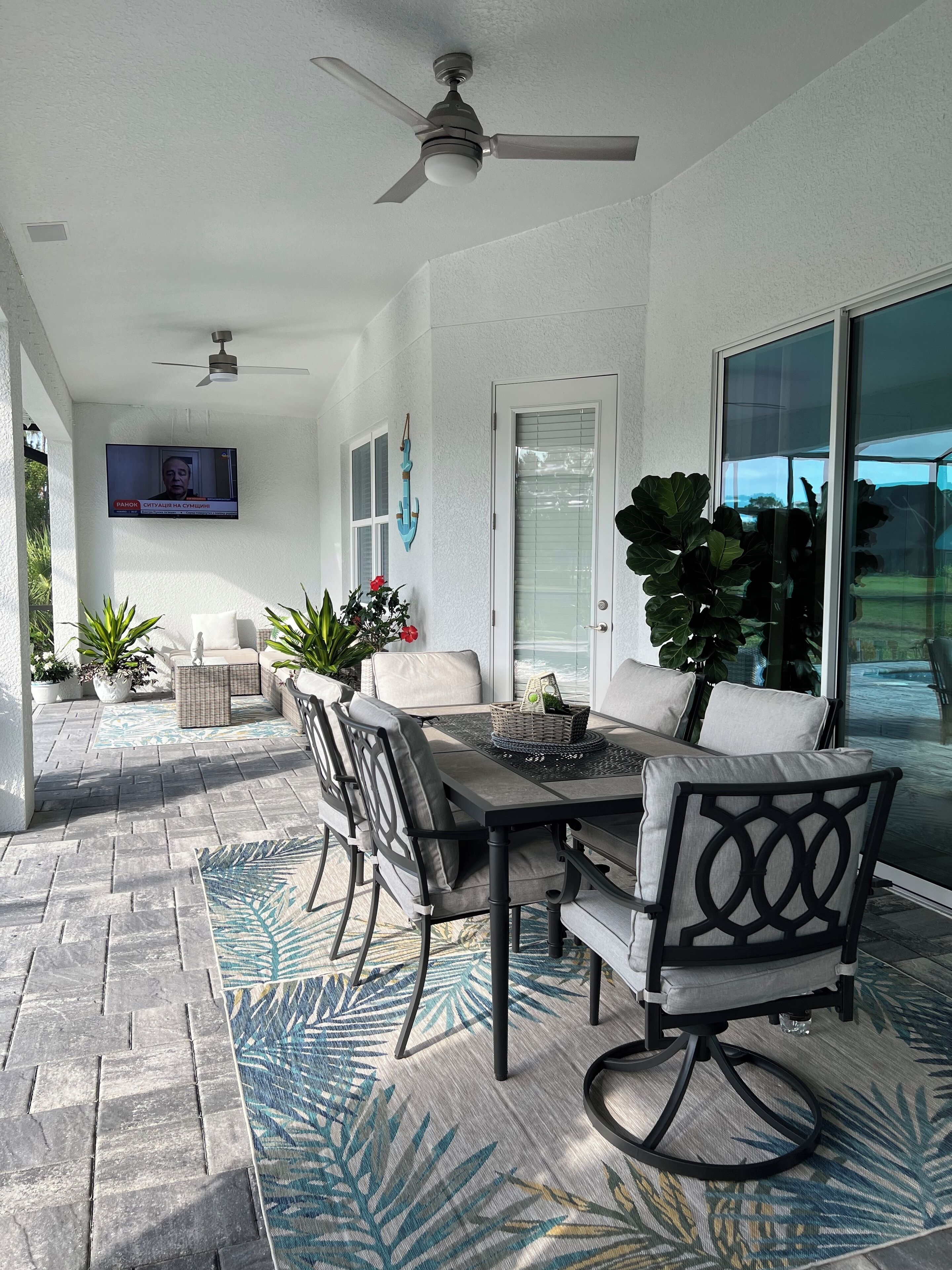 A covered patio area with a table and chairs, a ceiling fan, and a television screen displaying a news program.