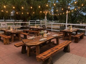 Un espace de restauration en plein air avec des tables et des bancs en bois rustique, des guirlandes lumineuses et une clôture blanche en arrière-plan.