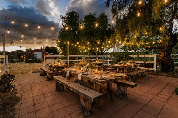 Un área de comedor al aire libre con mesas y bancos de madera, luces colgantes y una valla rústica.
