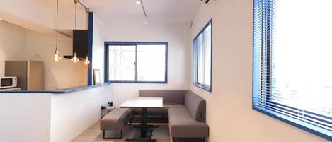 A modern kitchen with a microwave, a dining table, and a bench. There are two windows with blinds, and an air conditioning unit on the wall.