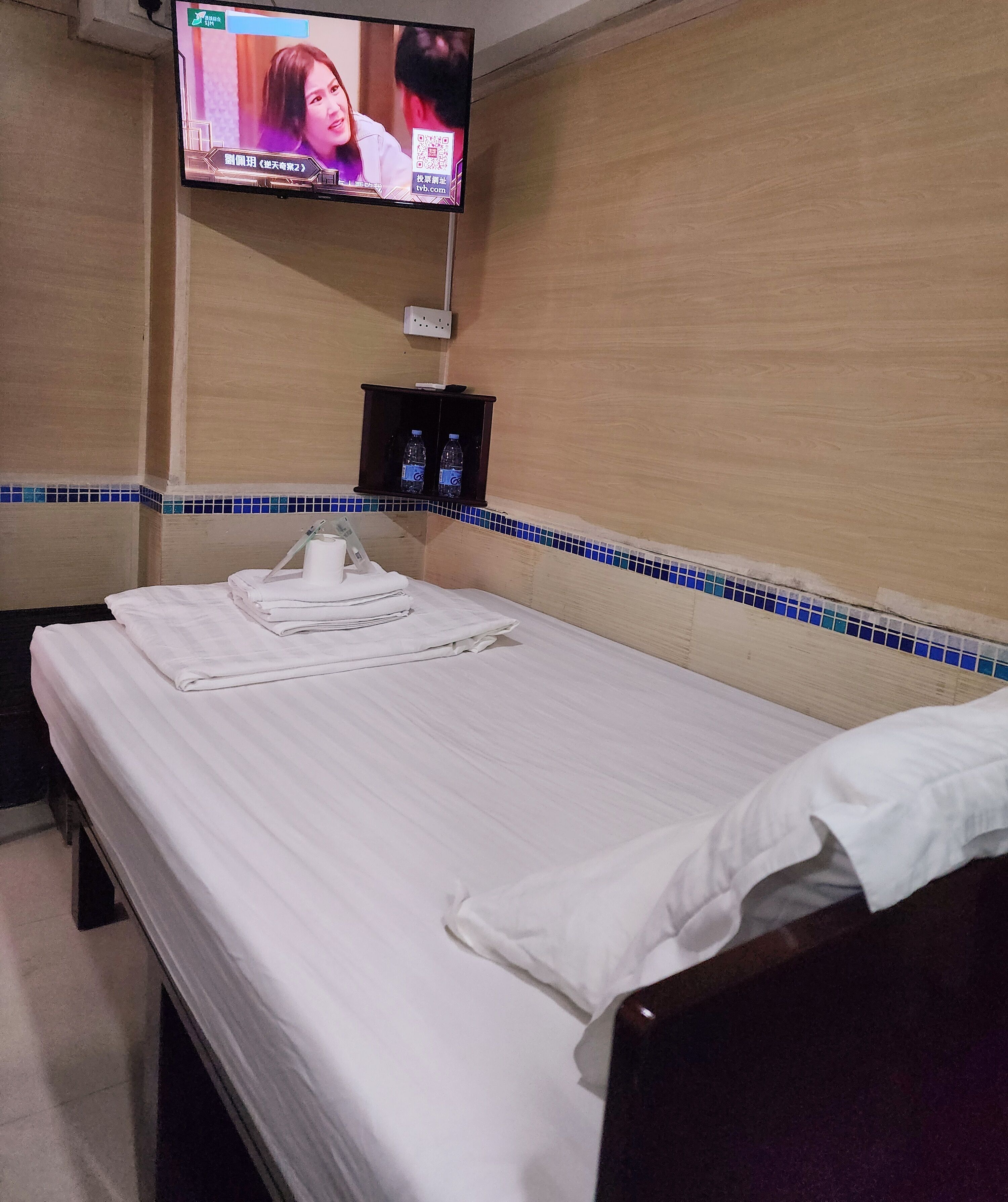 A hotel room with a bed, a TV mounted on the wall, and a small shelf with bottled water.