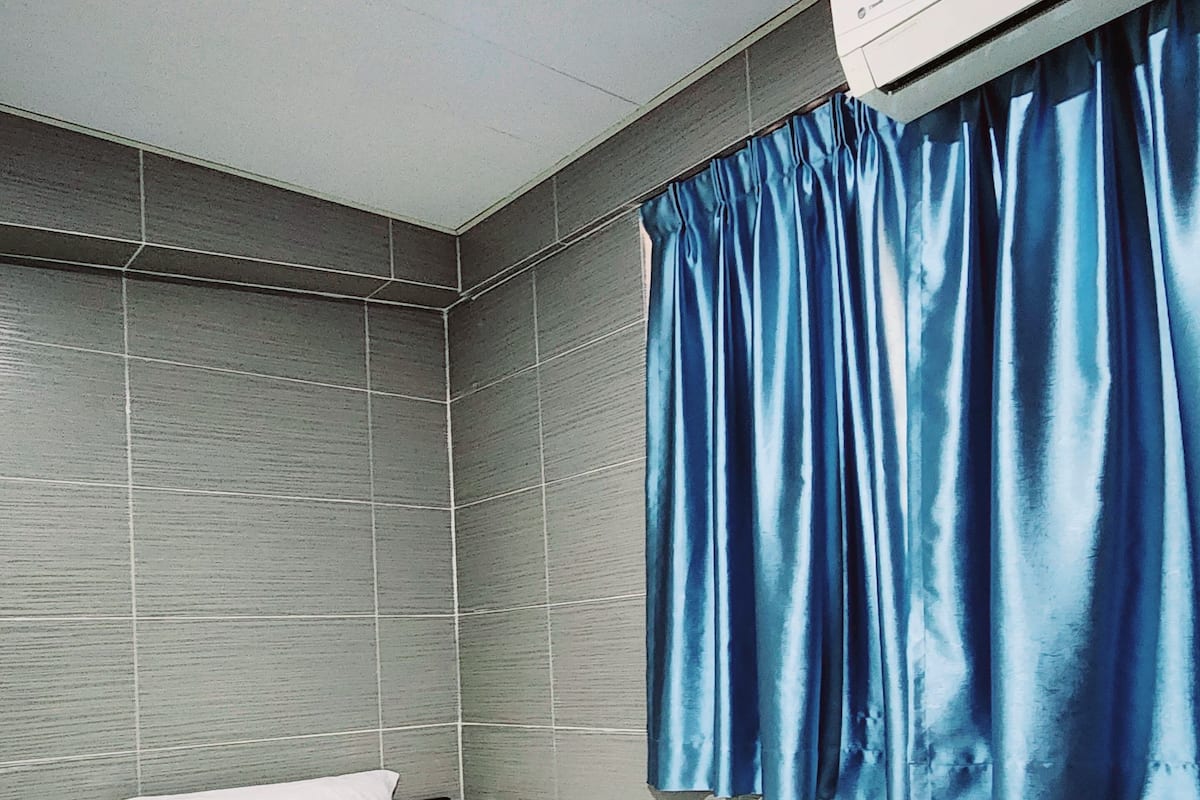 Une chambre d’hôtel avec un lit, une tête de lit, une fenêtre avec des rideaux bleus, un appareil de climatisation et une pile de serviettes sur le lit.