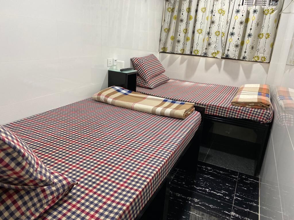 Ein kleiner, kompakter Raum mit zwei Betten, beide mit karierten Bettdecken. Ein kleiner Tisch mit Lampe und eine Steckdose sind an der Wand angebracht. Das Zimmer hat ein Fenster mit einem blumigen Vorhang.