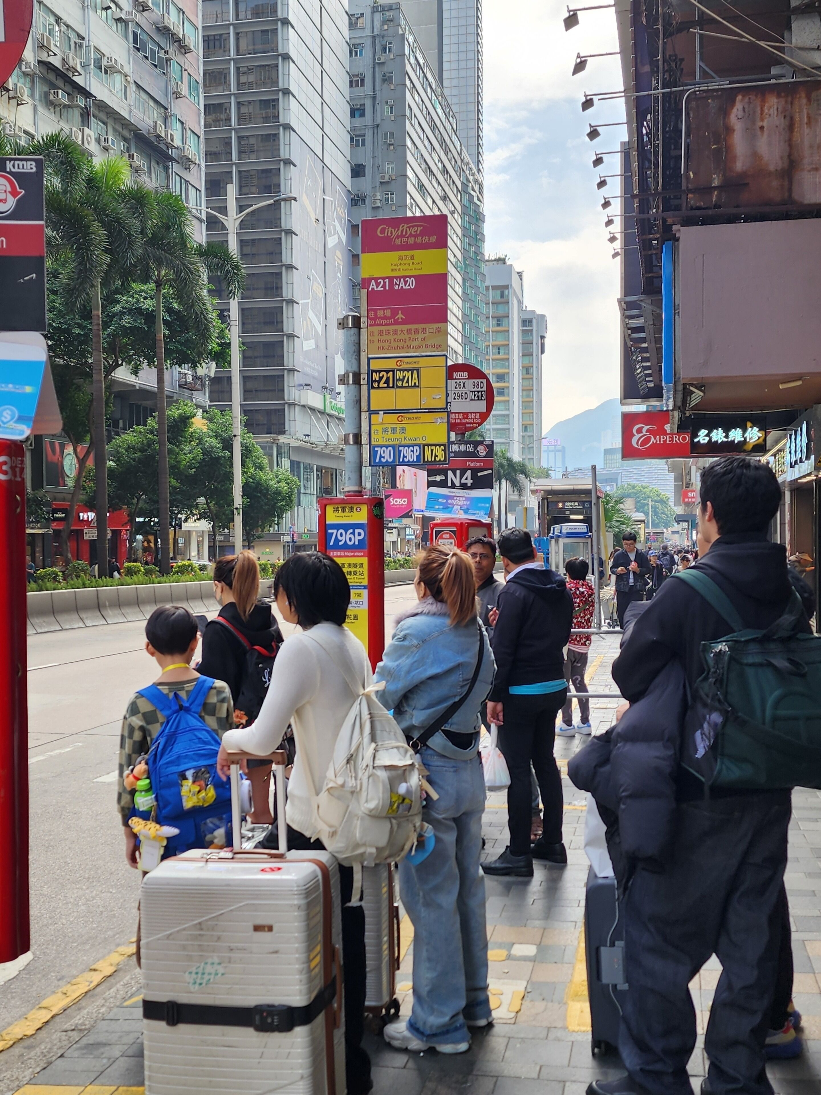 A busy urban street with people standing and walking, some with luggage.