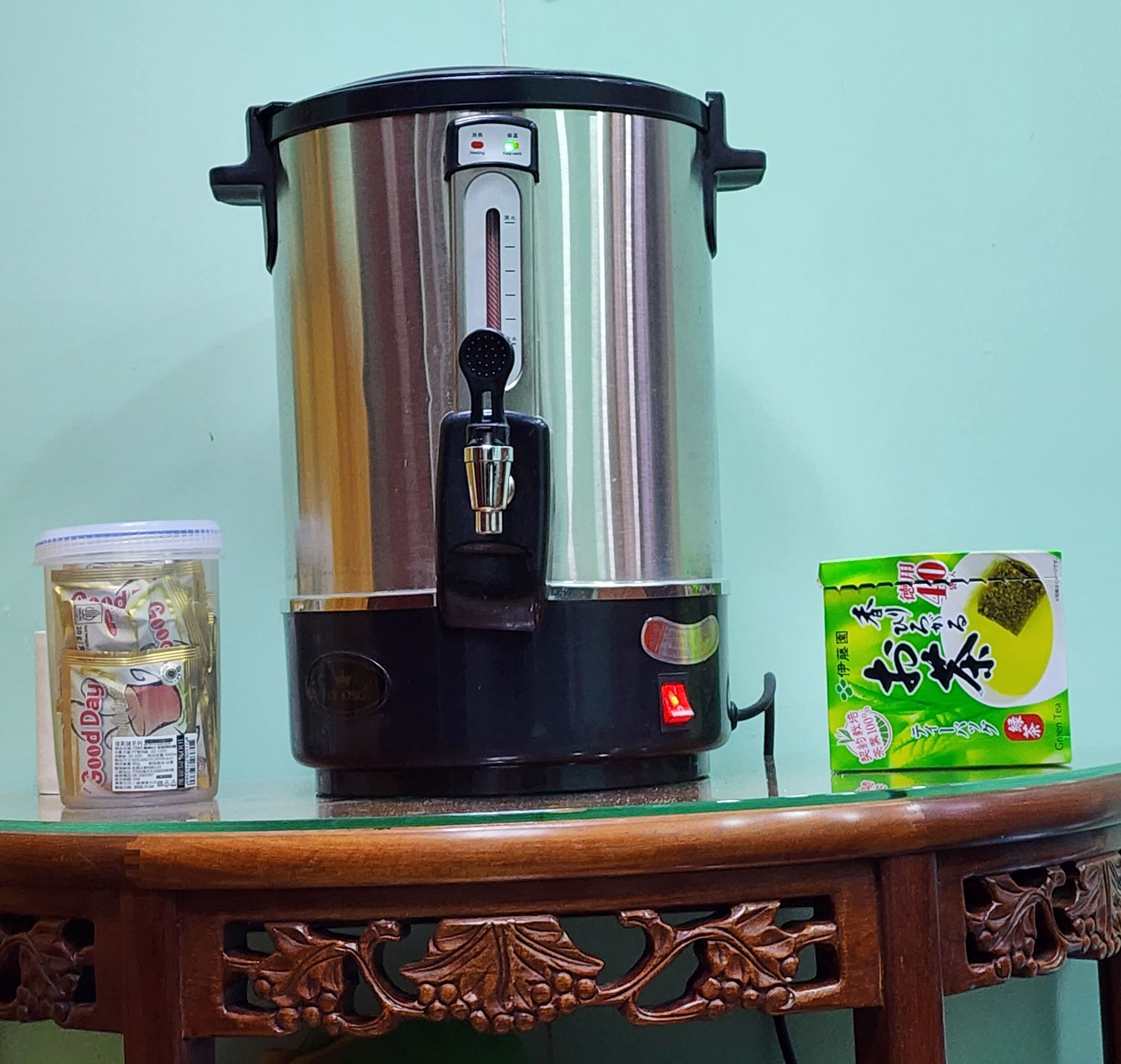 Une bouilloire électrique en acier inoxydable est posée sur une table en bois, à côté d’une boîte verte contenant du thé vert et d’un récipient en plastique rempli de café instantané.