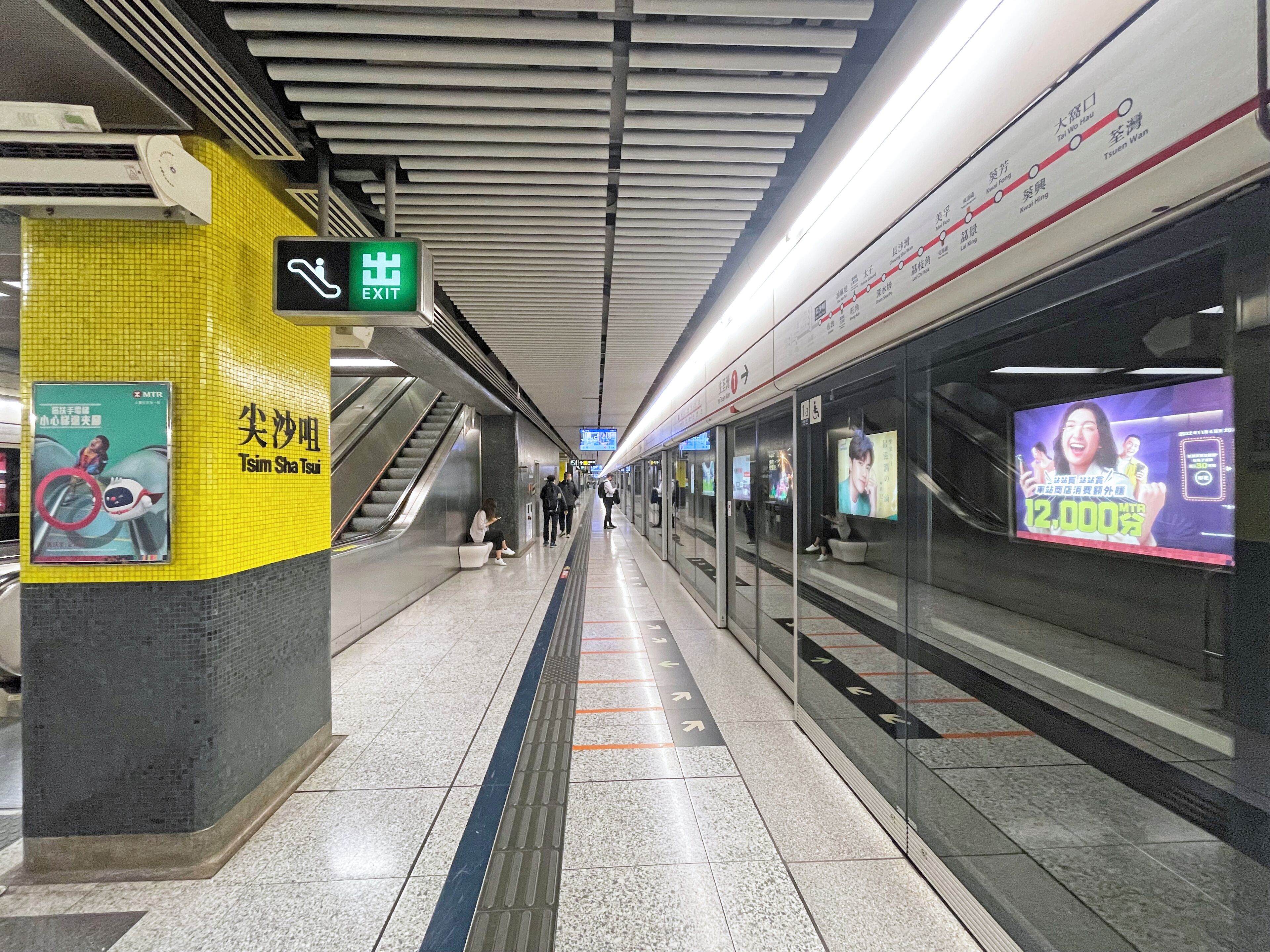 Estação de metrô com paredes revestidas de azulejos, placa de saída amarela e anúncios.