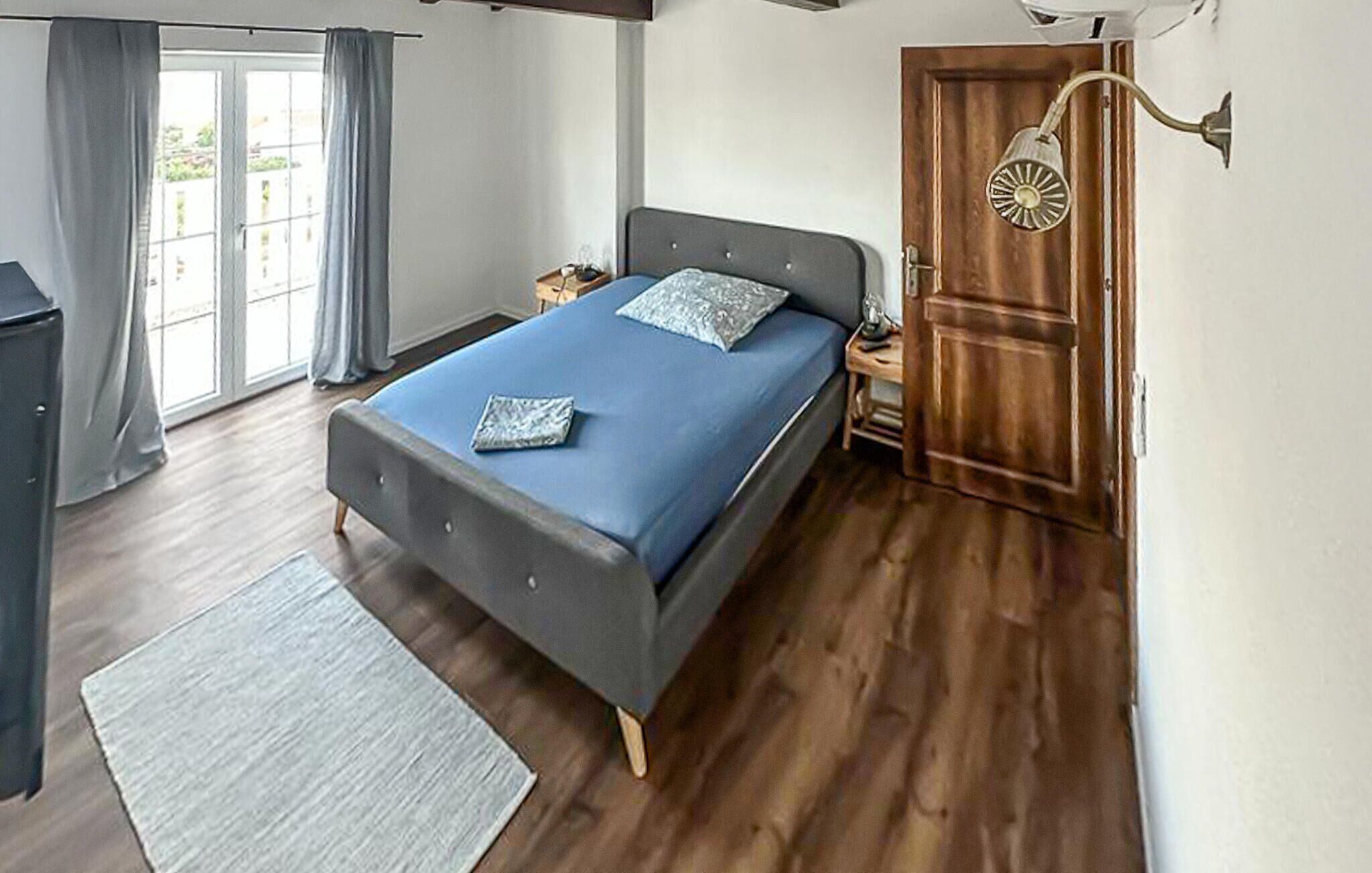 A bedroom with a bed, a nightstand, a lamp, a wooden door, and a window with curtains.