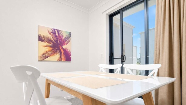 Une pièce moderne avec une table et des chaises blanches, une porte vitrée donnant sur l’extérieur et un tableau aux motifs tropicaux accroché au mur.