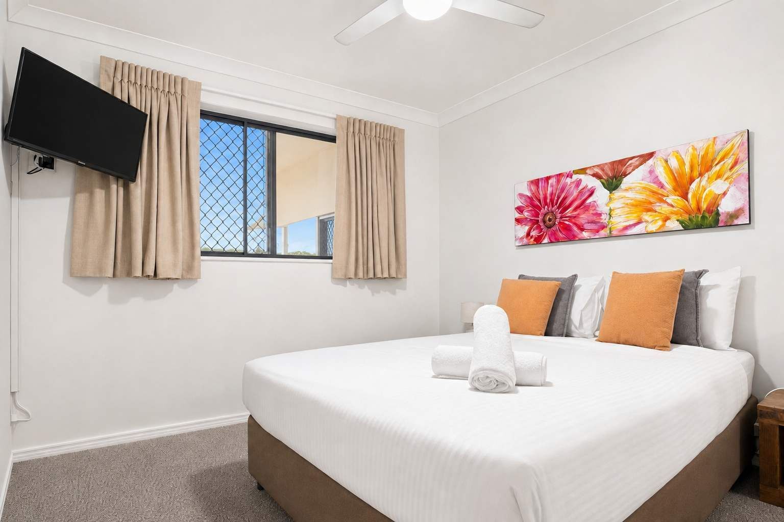 A hotel room with a bed, a flat-screen TV mounted on the wall, a window with curtains, and a floral artwork above the bed.