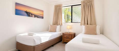 Quarto de hotel com duas camas, uma mesa de cabeceira, um ventilador de teto e uma janela com cortinas.
