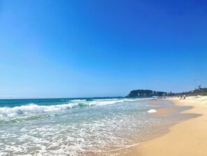 Ein Strand mit Wellen, blauem Himmel und Gebäuden im Hintergrund.