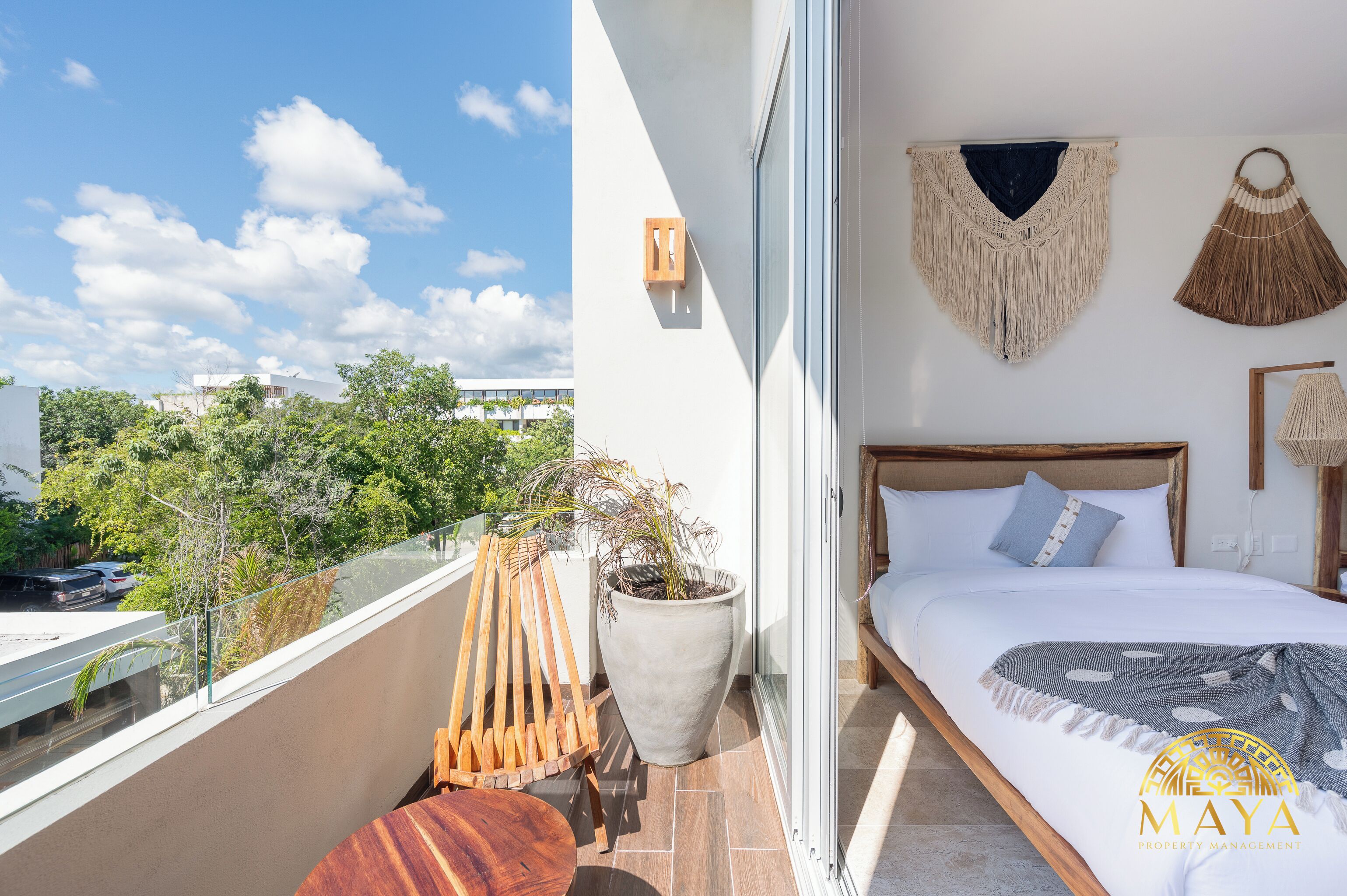 A balcony with a wooden chair and a potted plant, a bed with macrame wall hangings, and a view of a cityscape.