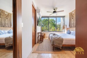 Un dormitorio moderno con dos camas, ventilador de techo, televisor y un ventanal con vistas a árboles.