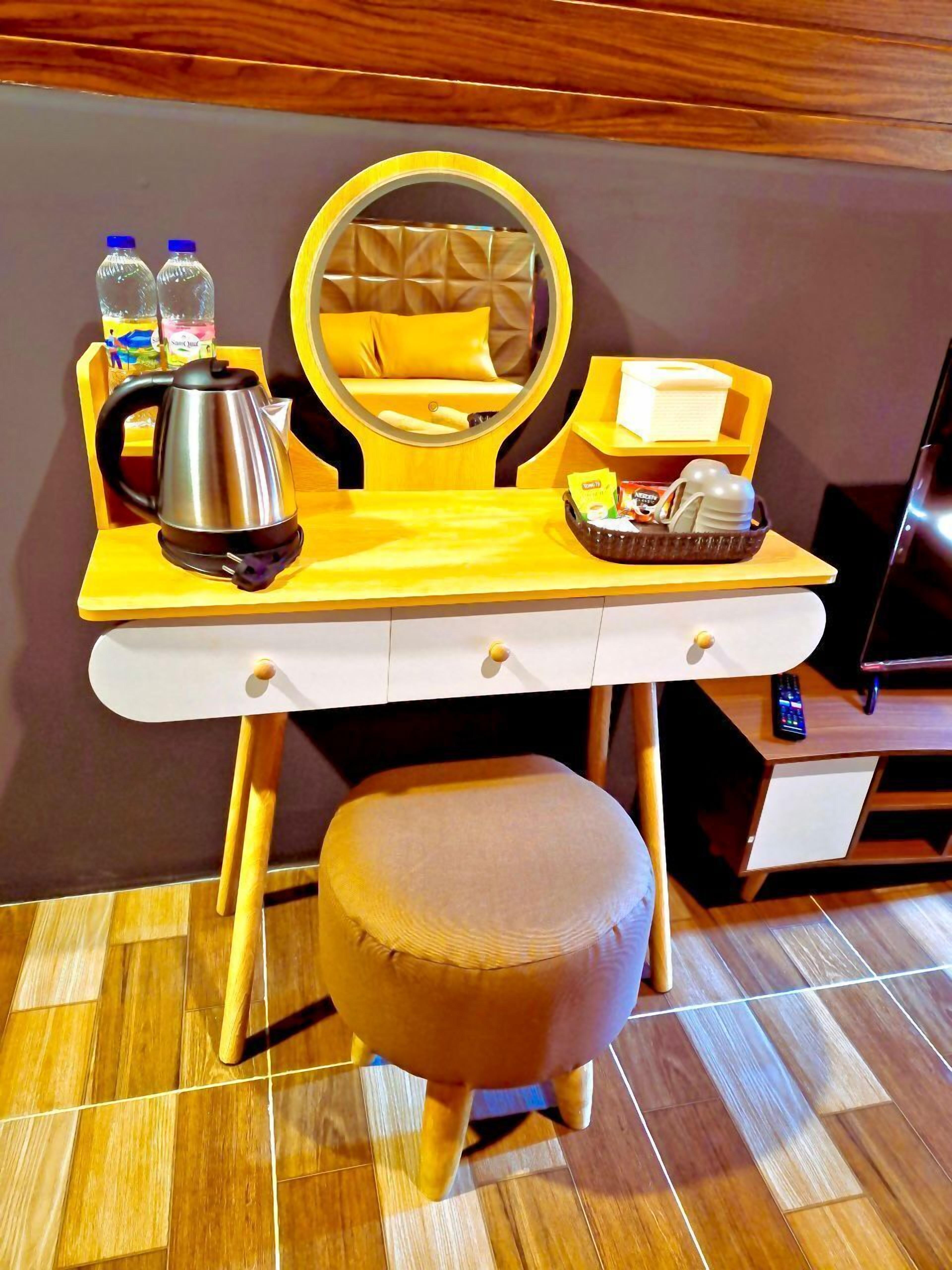 A compact hotel room with a wooden vanity, a round mirror, a small stool, a kettle, and a tray with toiletries.