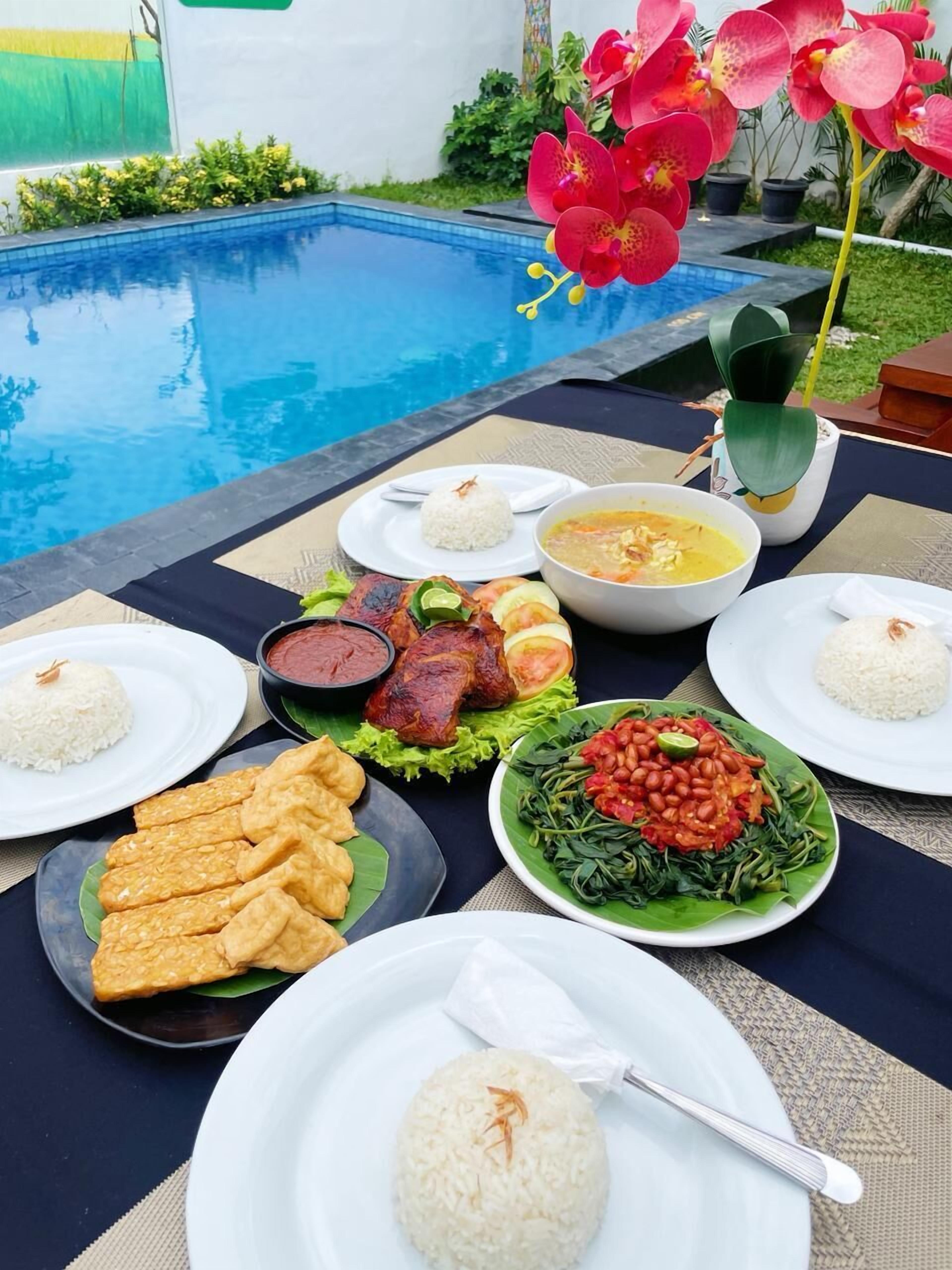 A dining table with various dishes including rice, soup, grilled meat, and vegetables, set against a poolside view.