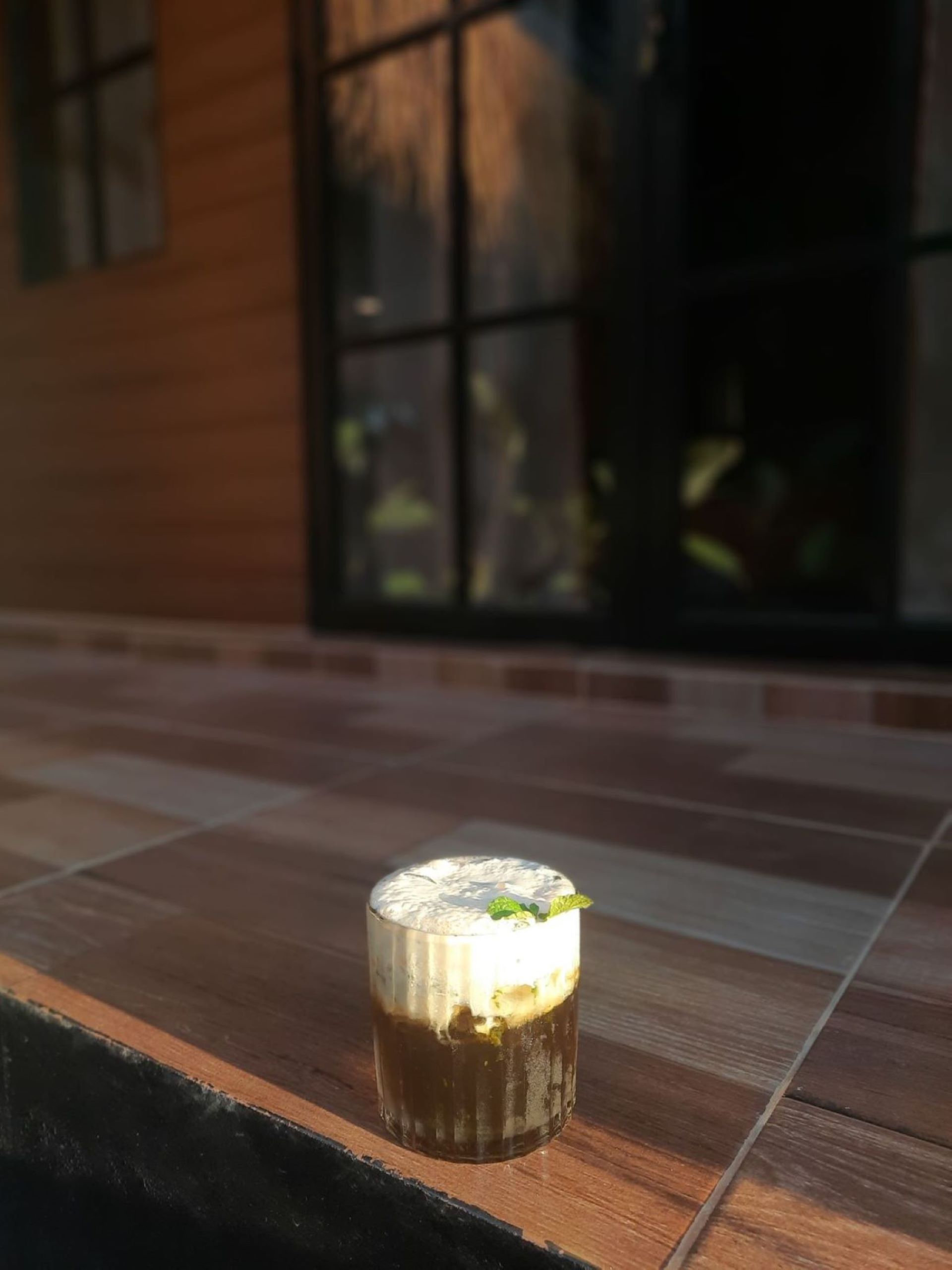 Un vaso de café helado sobre una mesa de madera con una ventana al fondo.