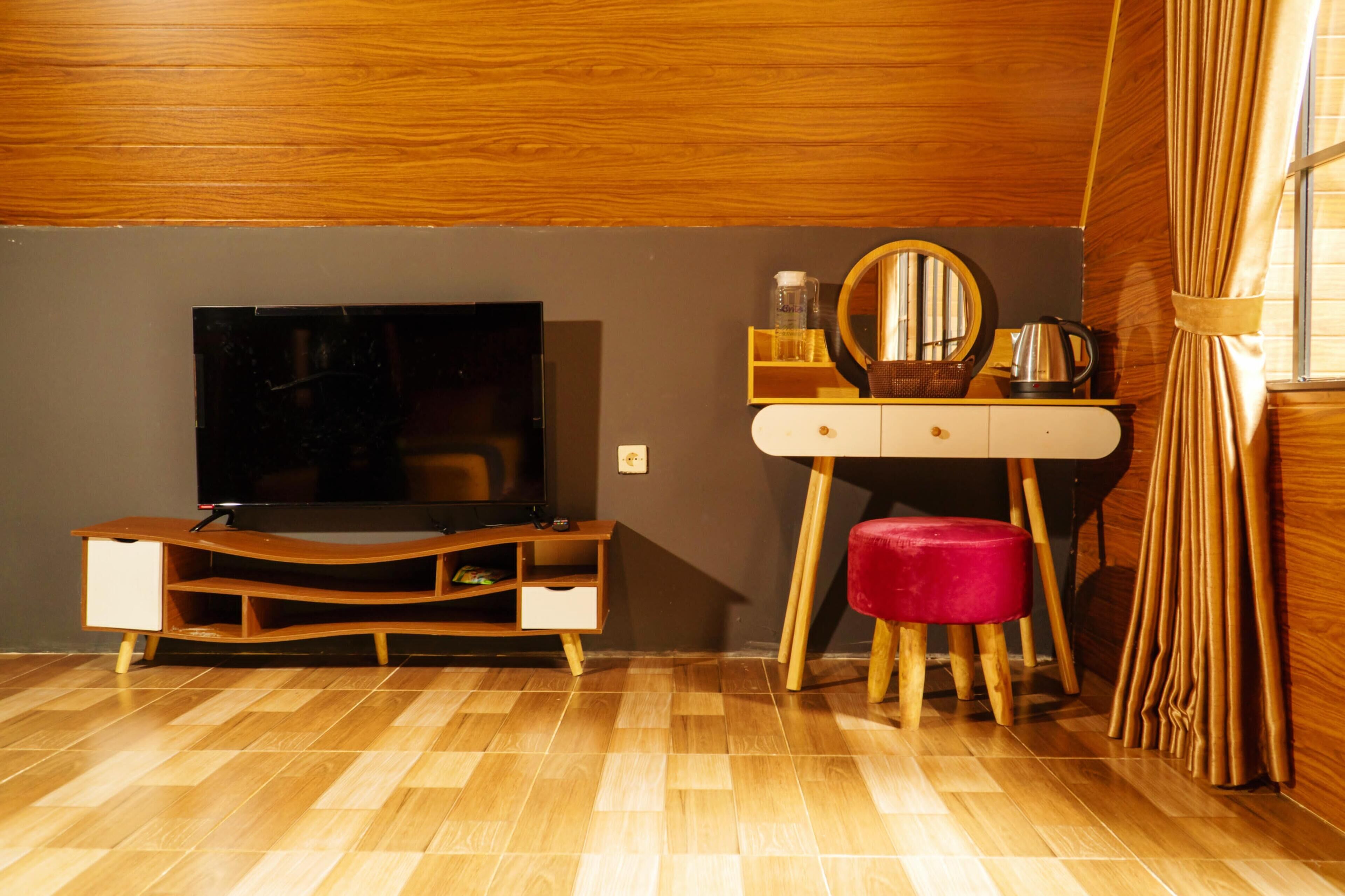 Une pièce comprenant un meuble de rangement en bois pour les appareils multimédias, un téléviseur, une coiffeuse avec un miroir et un tabouret rouge.