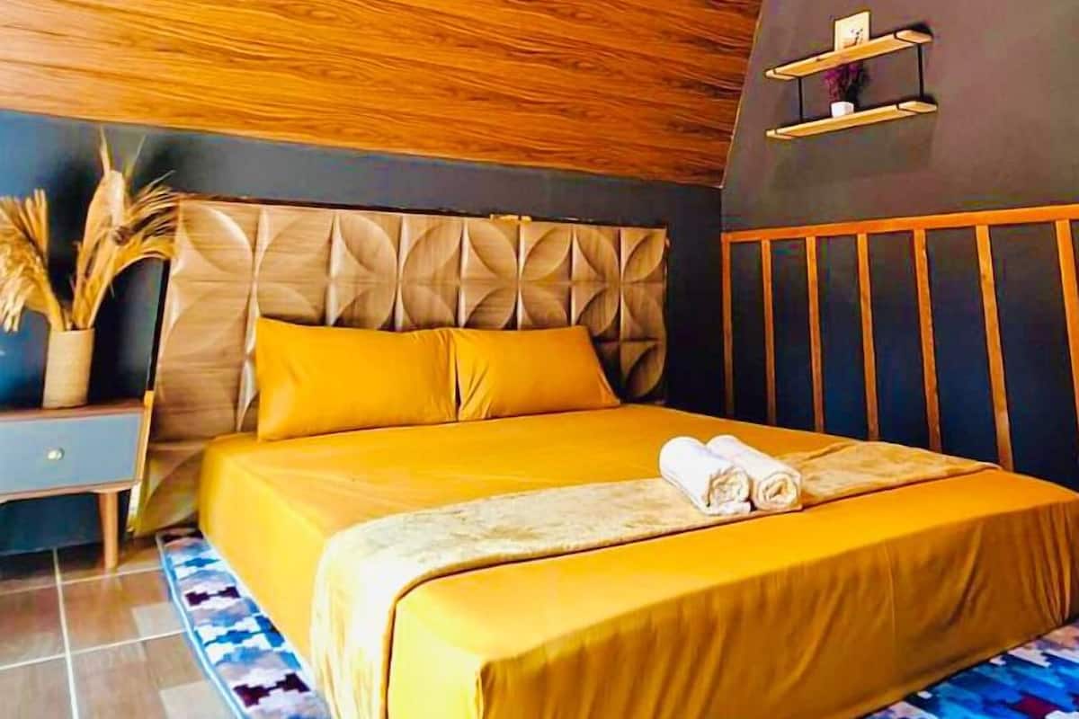 Un dormitorio moderno con techo de madera, una cama con ropa de cama amarilla, cabecera con diseño geométrico y una pequeña estantería con objetos decorativos.