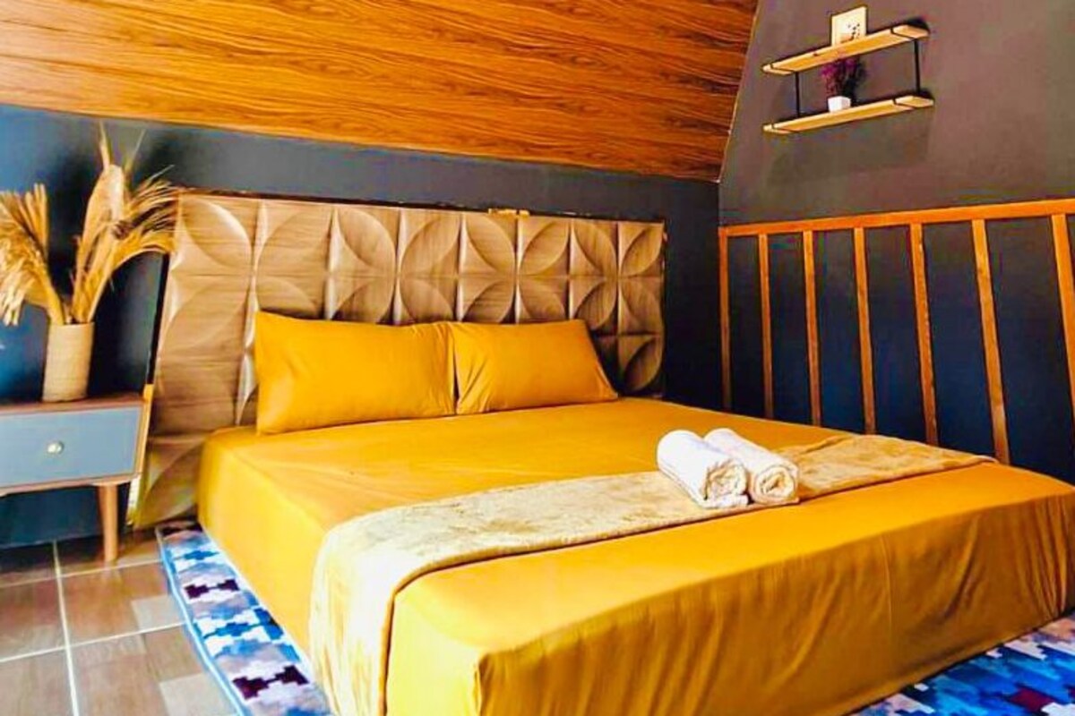 A modern bedroom with a wooden ceiling, a bed with yellow bedding, a bedside table, and a wall-mounted shelf.