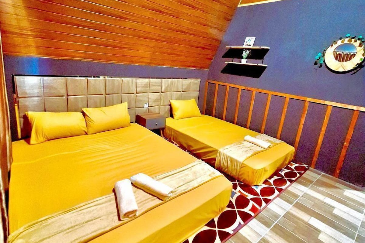 Habitación de hotel con techo de madera, una cama grande con ropa de cama amarilla, una cama más pequeña, una mesita de noche y un estante en la pared con un adorno.