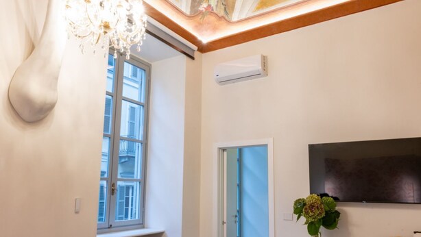 Una habitació amb un lampadari, una finestra amb persianes, un aparell d'aire condicionat, una porta i un televisió de pantalla plana penjada a la paret.